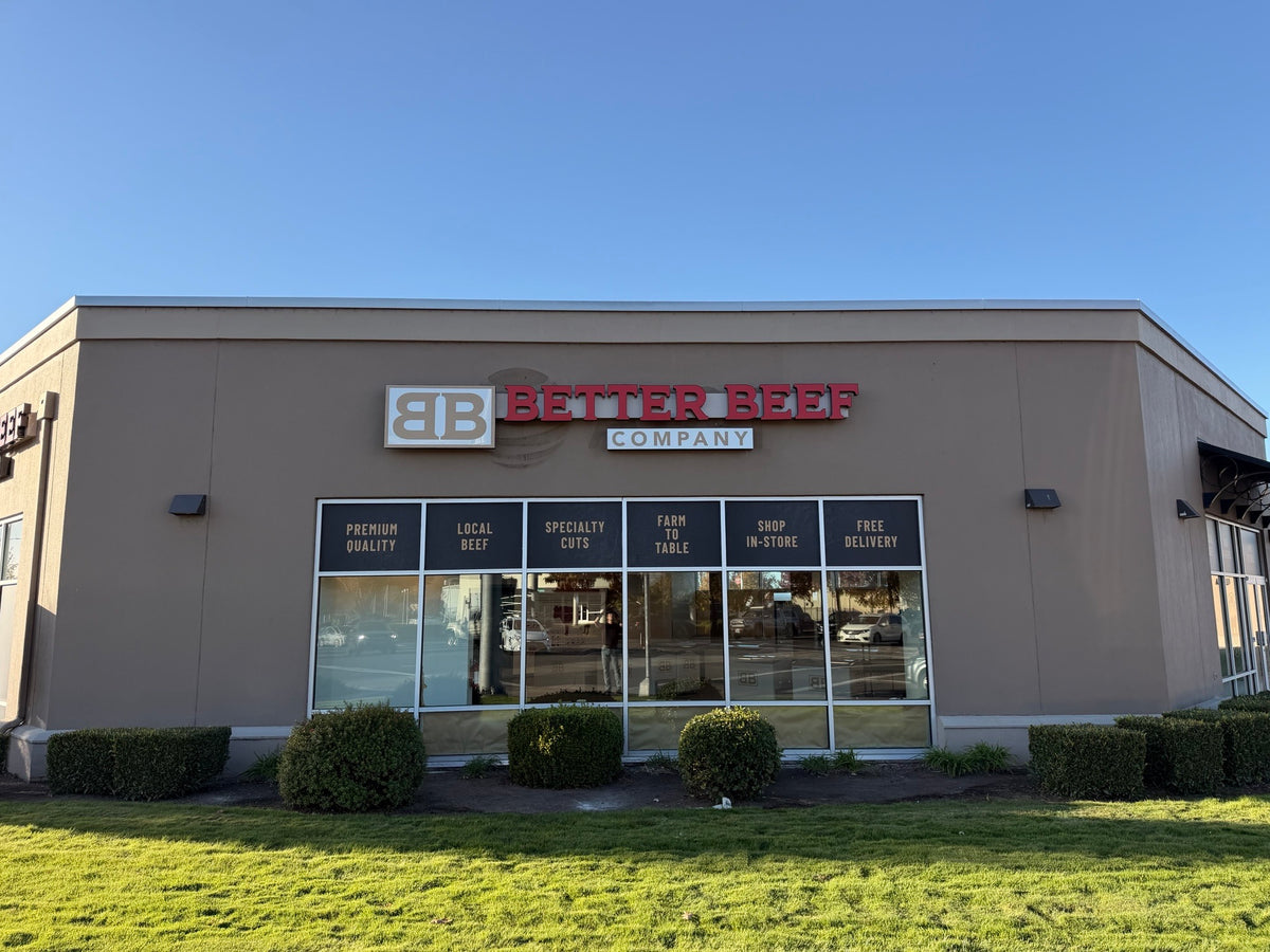 Front exterior of a Better Beef Company restaurant or retail building with a beige facade, large glass windows, red and white signage, grass, shrubs, and a clear blue sky.